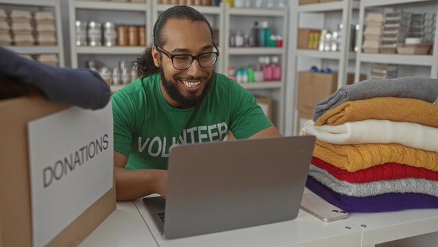 Man volunteer in green t shirt typing on laptop beside a labeled donations box and stacked sweaters in building; compassion community support. - Powered by Adobe