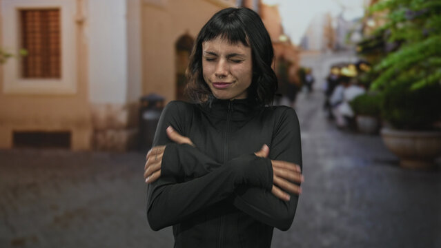 Young hispanic woman in black sportswear rubbing cold hands on a cobblestone street in winter dawn; determination. - Powered by Adobe