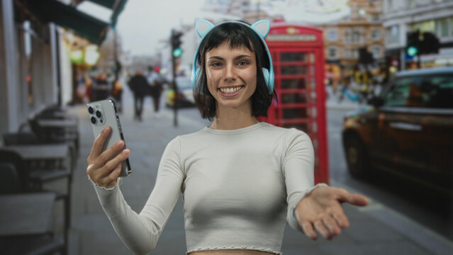 Young woman wearing crop top and cat ear headphones holds smartphone and beckons hand on street; invitation connection warmth.