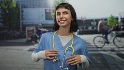 Woman nurse wearing blue scrubs adjusting yellow stethoscope around neck in a busy street with blurred cyclists and urban buildings; compassion.