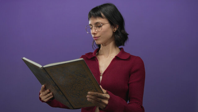 Woman smiling holding antique book with hands in purple studio wearing round glasses reading intently; curiosity.