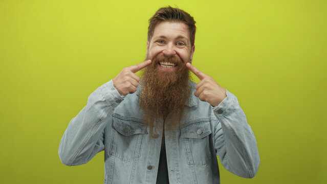 Man with long beard wearing denim jacket and black shirt points finger to teeth in a vibrant green studio; happiness.