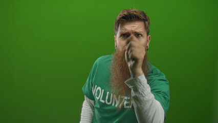 Man wearing a volunteer shirt with beard points finger at camera in a bright green studio set; urgency awareness activism motivation inspiration call signal alert concern drive.