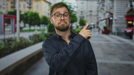 Man points finger upward on city street, wearing glasses and navy shirt, slight smile and raised eyebrow toward nearby building; amused curiosity.