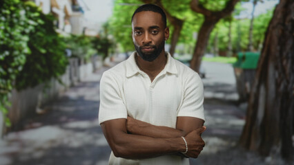 Fototapeta premium Man with arms crossed on a tree lined street looking downward in a white polo shirt wearing a beaded bracelet by a residential wall; contemplation.