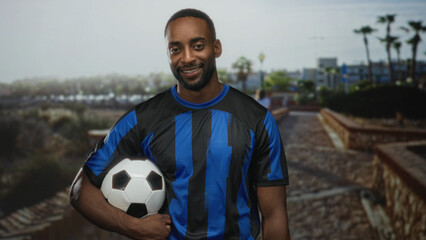 Man holding soccer ball under arm on street, smiling in blue striped jersey, young african american supporter; team pride.