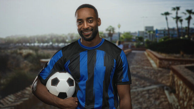 Man holding soccer ball under arm on street, smiling in blue striped jersey, young african american supporter; team pride. - Powered by Adobe