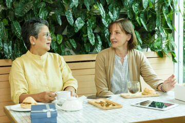 Asian senior woman and Caucasian woman sit at a cafe table, smiling warmly and engaging in a joyful conversation while enjoying tea, pastries, and a relaxed retirement moment., Concept small business