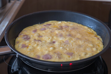 Close-up shot of an egg mixture with ham and onion pieces cooking in a black non-stick frying pan. The omelet is still partially liquid with visible bubbles