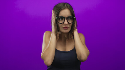 Woman holds both hands over ears wearing glasses and a navy tank top in studio setting with worried expression; stress.