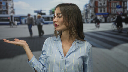 Young hispanic woman holds out hand on busy city street wearing glasses and striped blue shirt with...