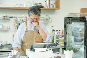 Asian senior female cafe owner wearing apron appearing thoughtful holding chin while looking at tablet managing receipts reflecting on small coffee shop business enjoying independent lifestyle.