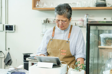 Asian senior female cafe owner concentrating using tablet standing at counter preparing orders confidently enjoying managing independent post-retirement coffee shop small family business.