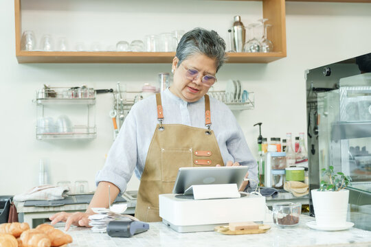 Asian senior female cafe owner standing at counter using tablet managing orders appearing calm enjoying post-retirement lifestyle running independent small coffee shop confidently.