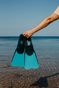A man's hand holds flippers against the sea.