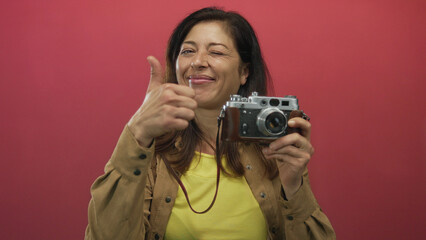Beaming friendly hispanic woman holding vintage camera gestures thumbs up in bright pink studio; approval.