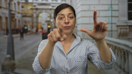 Middle aged hispanic woman points finger and shapes loser l gesture on busy city street with...