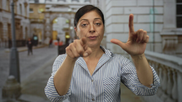 Middle aged hispanic woman points finger and shapes loser l gesture on busy city street with blurred passersby; mockery.