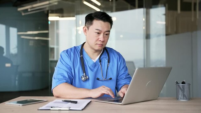 Confident asian doctor in blue coat typing on laptop sitting at workplace in hospital office. Focused handsome medical worker physician is engaged in work or chats online with patient using computer
