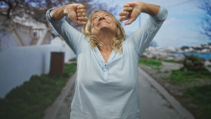 Senior woman with arms outstretched on a street, head tilted back and eyes closed, wearing light...
