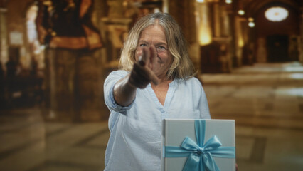 Senior woman points finger at wrapped gift box with blue ribbon inside ornate building; celebration joy.