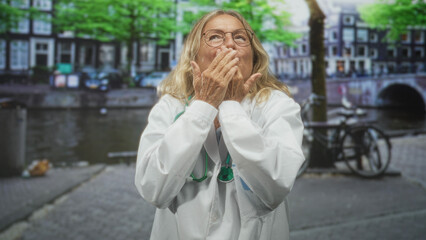 Fototapeta premium Woman doctor, senior and blonde, in a white labcoat with stethoscope blows a kiss with hands over mouth on a street by a canal; gratitude care warmth.