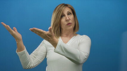 Woman in white top raises open palms and shrugs shoulders with puzzled expression in a front facing blue studio setting; uncertainty.