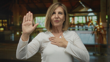 Middle age woman raising hand in hotel lobby building with palm forward and hand on chest; honor determination.