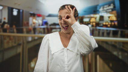 Obraz premium Young blonde woman smiling and making ok sign with hand to eye in a busy shopping mall building, wearing white blouse; joy discovery.