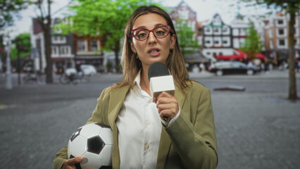 Woman reporter holds microphone and soccer ball while speaking on busy street; confidence sports reporting.