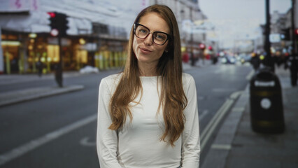 Obraz premium Woman with glasses smiling and gesturing with open hands on a busy city street near shops and traffic lights; friendly welcoming joy.