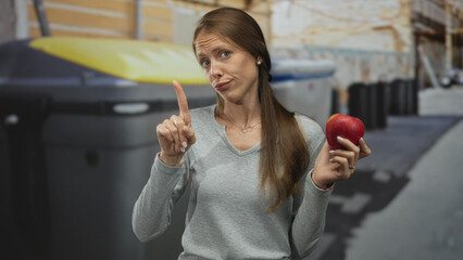 Woman holding apple and pointing finger on street near recycling and trash containers, showing...
