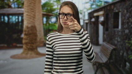 Woman wearing striped top and glasses points finger at camera in stone building courtyard with bench and tree trunk visible; confidence instruction.