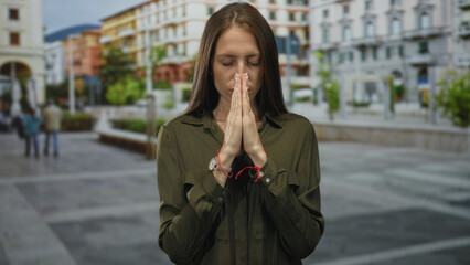 Fototapeta premium Woman with hands pressed together in prayer gesture, closed eyes and visible hands wearing a watch on a city street plaza; quiet contemplation.