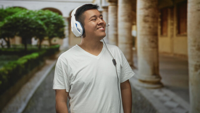 Hispanic young man wearing white headset with microphone, smiling and looking up in historic building corridor; serenity listening. - Powered by Adobe