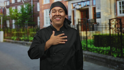 Hispanic chef man wearing black jacket with hand on chest at building entrance on street, smiling...