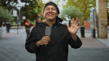 Man chef in black uniform smiling holding metal spatula and waving hand on street; welcome warmth...