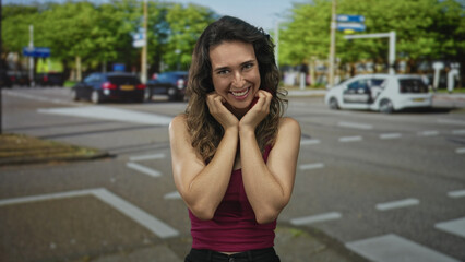 Fototapeta premium Young hispanic woman pressing cheeks with hands in a broad grin under sunlit trees on a busy street crosswalk; joy.