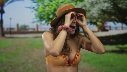 Woman in swimwear smiling playfully with hands mimicking binoculars, enjoying a sunny day in a...