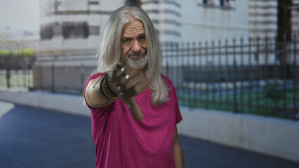Fototapeta premium Man with long gray hair and beard showing thumbs down gesture on street in front of building fence and railing, wearing pink t shirt; disapproval defiance.