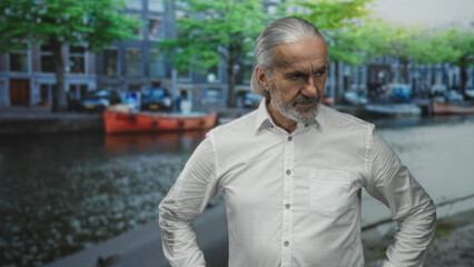 Man middle age with long gray hair standing with hands on hips on a street by a canal in amsterdam...