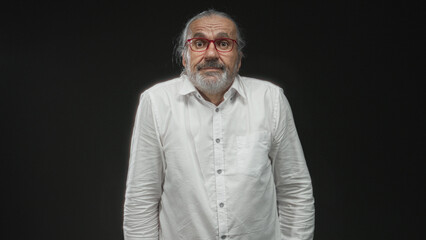 Middle aged man with hoary long hair and red glasses shrugs shoulders in studio against black wall; uncertainty reflection.