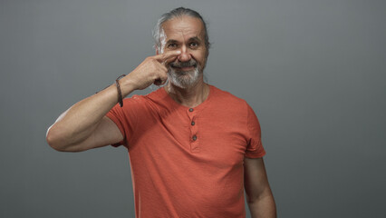 Man with hoary long hair wearing orange shirt points finger to nose in gray studio; playful mischief.