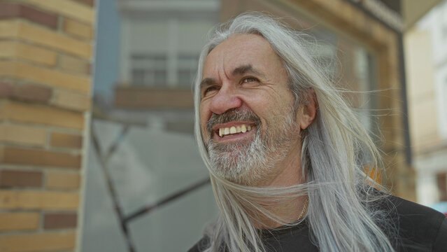 Man with long silver hair and full beard smiling as wind lifts strands across his face on a street by a brick building; joy wisdom.