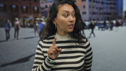 Woman holds up index finger on a busy city street while raising an eyebrow and glancing sideways;...