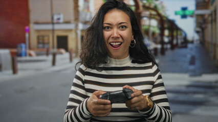 Woman holding a game controller with both hands, thumbs pressing buttons, standing on a city street near shopfronts; playful joy.