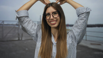 Smiling woman wearing glasses makes heart shape with hands on street pier by ocean walkway; love joy.