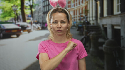 Young blonde woman in pink t shirt wearing bunny ears extends closed fist for bump on cobblestone street; friendship.