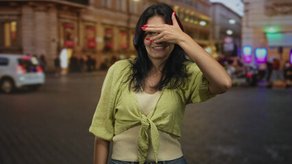 Fototapeta premium Woman covers eyes with a hand gesture in front of a lit building on street crowded with pedestrians; playfulness.