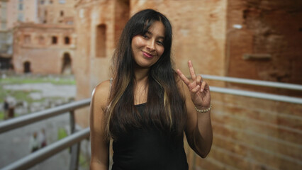 Brunette woman with long hair in black tank top holds up peace sign by railing at roman ruins...